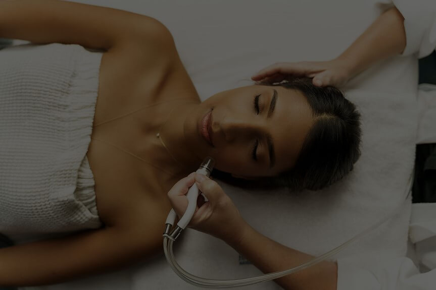 Person lying down with eyes closed and wrapped in a towel, receiving a facial treatment.